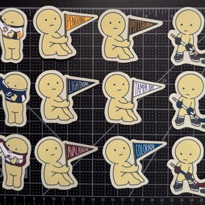 May include: A collection of twelve die-cut stickers featuring a pale yellow cartoon character in various hockey-themed poses, including wearing jerseys, holding pennants labeled Penguins, Pittsburgh, Lightning, Tampa Bay, Avalanche, and Colorado.