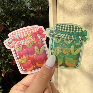Op de afbeelding: Twee stickers met potten fruitconserven. E&eacute;n pot is roze met rode appels, de andere is groen met groene appels. Elke pot heeft een geruite deksel, een strik en een holografisch effect.
