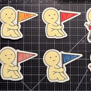 May include: A collection of eight die-cut stickers featuring pale yellow cartoon figures. Some figures hold pennants labeled Flames, Calgary, Goldeneyes, or Vancouver. Others wear jerseys or hold hockey sticks with pucks on a black cutting mat.