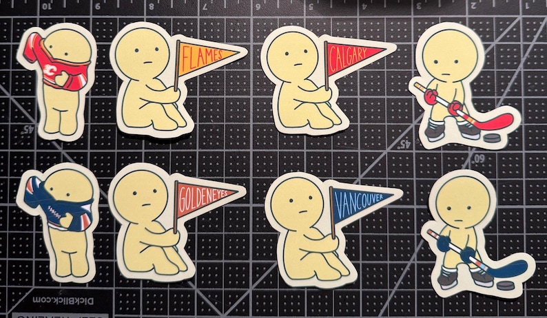 May include: A collection of eight die-cut stickers featuring pale yellow cartoon figures. Some figures hold pennants labeled Flames, Calgary, Goldeneyes, or Vancouver. Others wear jerseys or hold hockey sticks with pucks on a black cutting mat.