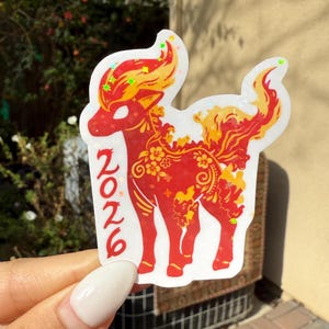 May include: A sticker depicting a red deer-like figure with yellow and orange details, including floral patterns. The number "2026" is in red. The sticker is held up by a hand.