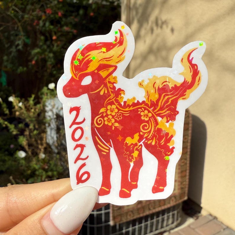 May include: A sticker depicting a red deer-like figure with yellow and orange details, including floral patterns. The number "2026" is in red. The sticker is held up by a hand.