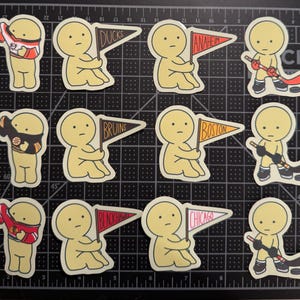 May include: A collection of twelve die-cut stickers featuring yellow cartoon figures. The stickers depict figures wearing hockey jerseys, holding pennants labeled Ducks, Anaheim, Bruins, Boston, Blackhawks, and Chicago, or playing hockey with sticks and pucks.