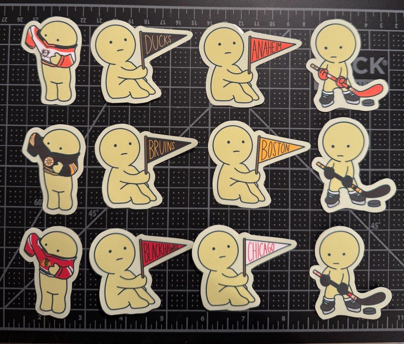 May include: A collection of twelve die-cut stickers featuring yellow cartoon figures. The stickers depict figures wearing hockey jerseys, holding pennants labeled Ducks, Anaheim, Bruins, Boston, Blackhawks, and Chicago, or playing hockey with sticks and pucks.