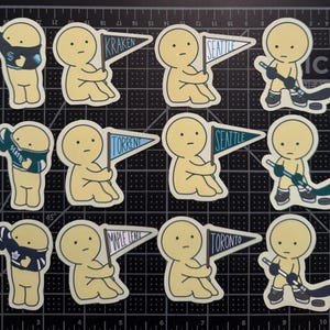 May include: A collection of twelve die-cut stickers featuring pale yellow cartoon figures. Some figures wear sports jerseys, some hold pennant flags labeled Kraken, Seattle, Torrent, Maple Leafs, or Toronto, and others hold hockey sticks with pucks.