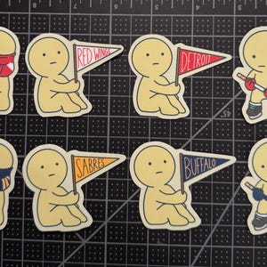 May include: A set of eight die-cut stickers featuring cartoon figures in various poses. Four figures hold pennants labeled Red Wings, Detroit, Sabres, and Buffalo. Two figures wear hockey jerseys, and two figures hold hockey sticks with pucks.