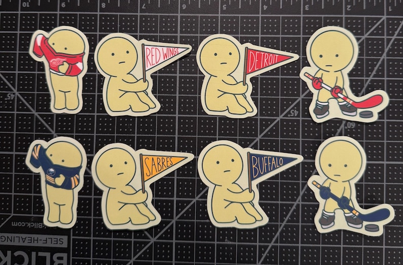 May include: A set of eight die-cut stickers featuring cartoon figures in various poses. Four figures hold pennants labeled Red Wings, Detroit, Sabres, and Buffalo. Two figures wear hockey jerseys, and two figures hold hockey sticks with pucks.