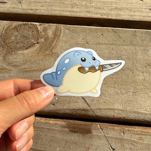 Puede incluir: Una pegatina troquelada que representa una criatura de dibujos animados con un cuerpo azul claro, vientre crema y boca marr&oacute;n mordiendo un cuchillo plateado. La criatura tiene ojos negros y reflejos blancos. La pegatina se sostiene sobre un fondo de madera.
