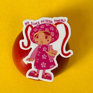 May include: A pink and white sticker with a cartoon girl wearing a pink hat with a flower pattern and a pink dress with a flower pattern. The sticker says "No super pattern powers?"