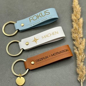 Könnte beinhalten: Drei Schlüsselanhänger aus Leder mit goldfarbenen Metallringen. Der hellblaue trägt die Aufschrift FOKUS 1% BESSER JEDEN TAG, der weiße zeigt einen Stern und das Wort MACHEN, der braune ist mit DISZIPLIN > MOTIVATION graviert.