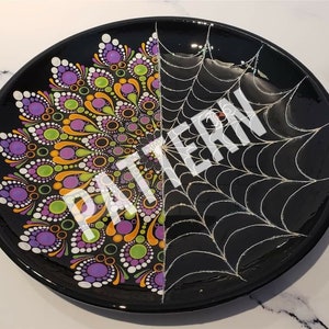Może przedstawiać: Czarny talerz dekoracyjny z połową wzoru mandali w fioletowe, zielone, pomarańczowe i białe kropki oraz wzorem białej pajęczyny na drugiej połowie. Słowo "PATTERN" jest napisane na talerzu na biało.