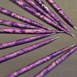 May include: A collection of nail art dotting tools featuring a marbled purple and violet finish with gold metallic flecks. Each tool has a tapered cylindrical handle and a thin, pointed metal tip extending from one end for precision work.