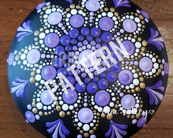 May include: A purple and gold mandala painted on a black round stone. The mandala features a circular pattern of dots in various shades of purple and gold.