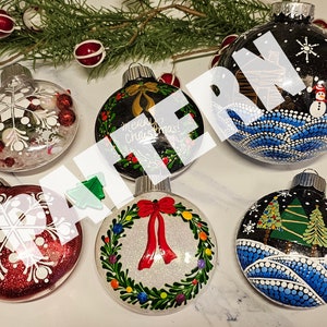 May include: Six glass Christmas baubles with various painted designs. One bauble features a snowman and a cabin, another has a wreath with a red bow, and another has a snowflake design. The baubles are all painted with white, red, green, and blue paint.