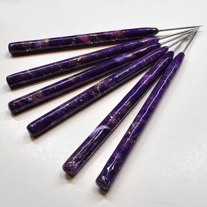 May include: A set of five crochet hooks with smooth, cylindrical handles in a deep purple marbled pattern featuring embedded gold-colored flecks. Each handle is attached to a slender, pointed silver-colored metal hook tip for needlework.