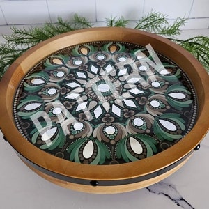 May include: A round wooden tray with a black metal rim and a green, brown, and white mandala design painted on the inside.
