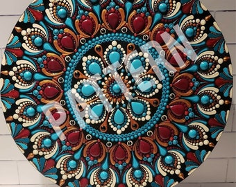 May include: A round mandala painted on a wooden surface with a black background. The mandala features a circular pattern of turquoise, red, gold, and white dots. The mandala is on a black stand.