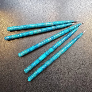 May include: A set of five long, slender tools with a marbled turquoise and blue finish. Each tool features a cylindrical body that tapers to a fine, sharp metal point at one end. The surface has a glossy, swirling pattern of light and dark blue.