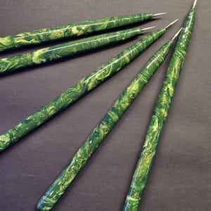 May include: A set of five slender tools with pointed metal tips and cylindrical handles featuring a marbled pattern of dark green, light green, and gold flecks. The handles have a smooth, glossy finish and taper toward the sharp metal points.