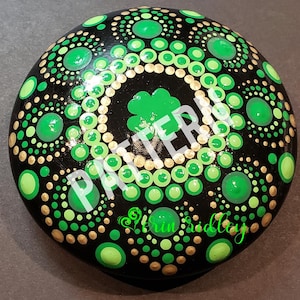 May include: A round, black stone painted with a green shamrock in the center. The shamrock is surrounded by concentric circles of green and gold dots.