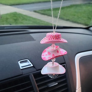 May include: A car accessory featuring three miniature pink cowboy hats hanging from a string. The top hat is covered in rhinestones, the middle hat has a cow print, and the bottom hat has a disco ball.