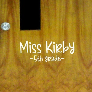 May include: A wooden door with a small window and a sign that reads "Miss Kirb - 5th grade - "