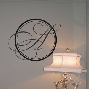 May include: A black and white monogram decal of the letter "A" in a decorative script font, inside a circle, on a light grey wall. A white lamp with a white lampshade is on a table below the decal.