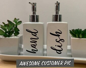 May include: Two white ceramic soap dispensers with black lettering. The dispenser on the left says "hand" and the dispenser on the right says "dish". The dispensers are sitting on a white tray with two small potted plants.