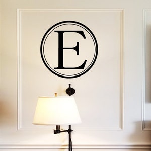 May include: A white bedroom with a black and white framed picture of a building with the text "Maison de Rue" on the wall. There is a black lamp with a white lampshade on a wooden table. The bed has a white headboard and white bedding with a gray and white patterned pillow.