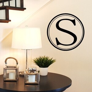 May include: A black and white monogram decal featuring the letter "S" within a circle. The decal is positioned on a white wall adjacent to a wooden staircase and a small table with a lamp and candles.
