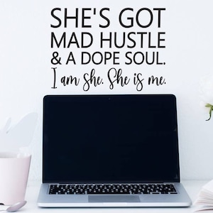 May include: A white wall with a black vinyl decal that reads "SHE'S GOT MAD HUSTLE & A DOPE SOUL. I am She. She is me.". A laptop, a pink mug, a book, a pen, and a vase of pink and white flowers are on a white surface in front of the wall.