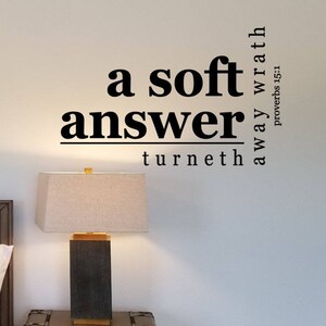 May include: A white wall with a black and white quote that reads "a soft answer turneth away wrath Proverbs 15:1". The wall is decorated with a brown woven mirror and a white lamp with a beige lampshade.