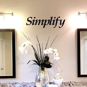 May include: A bathroom interior with two framed mirrors, a light fixture above each mirror, and a vase of white orchids on the counter. The word "Simplify" is written on the wall above the orchids.