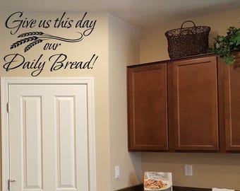May include: A kitchen scene featuring a white door and dark brown cabinets. Above the door, black script reads "Give us this day our Daily Bread!" A woven basket and greenery sit atop the cabinets. The wall is painted a light beige.