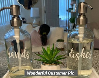 May include: Two clear glass bottles with silver pumps and labels that say "hands" and "dishes". The bottles are sitting on a white marble tray with a small green succulent plant in a white pot.
