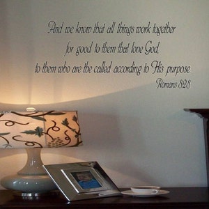 May include: A white wall with a black and white inspirational quote from the Bible, Romans 8:28, that reads "And we know that all things work together for good to them that love God, to them who are the called according to His purpose." The quote is displayed above a bedside table with a lamp and a picture frame.