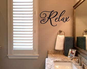 May include: A bathroom with white walls and a white window with white blinds. The bathroom has a large mirror with a silver frame. The mirror is above a white countertop with a sink and faucet. There is a towel rack with a white towel on it. The wall has a black vinyl decal that says "Relax".
