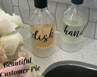 May include: Two clear glass pump bottles with white bases and black pumps. The bottles are labeled "dish" and "hand" in black script. The bottles are sitting on a white countertop next to a sink.