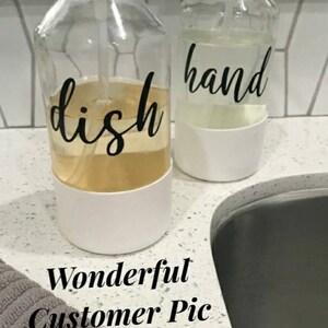 May include: Two clear glass pump bottles with white labels. The bottle on the left says "dish" and the bottle on the right says "hand". The bottles are sitting on a white countertop next to a stainless steel sink.