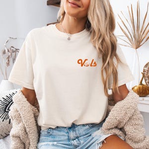 May include: A woman wearing a white t-shirt with the word "Vols" in orange lettering and a football graphic. She is also wearing blue denim shorts and a brown cardigan.