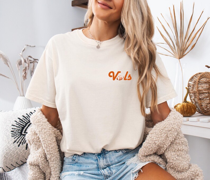 May include: A woman wearing a white t-shirt with the word "Vols" in orange lettering and a football graphic. She is also wearing blue denim shorts and a brown cardigan.