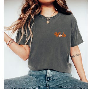 May include: A dark gray t-shirt with the word "Vols" in orange lettering and a small orange football graphic. The shirt is worn by a person with long brown hair.