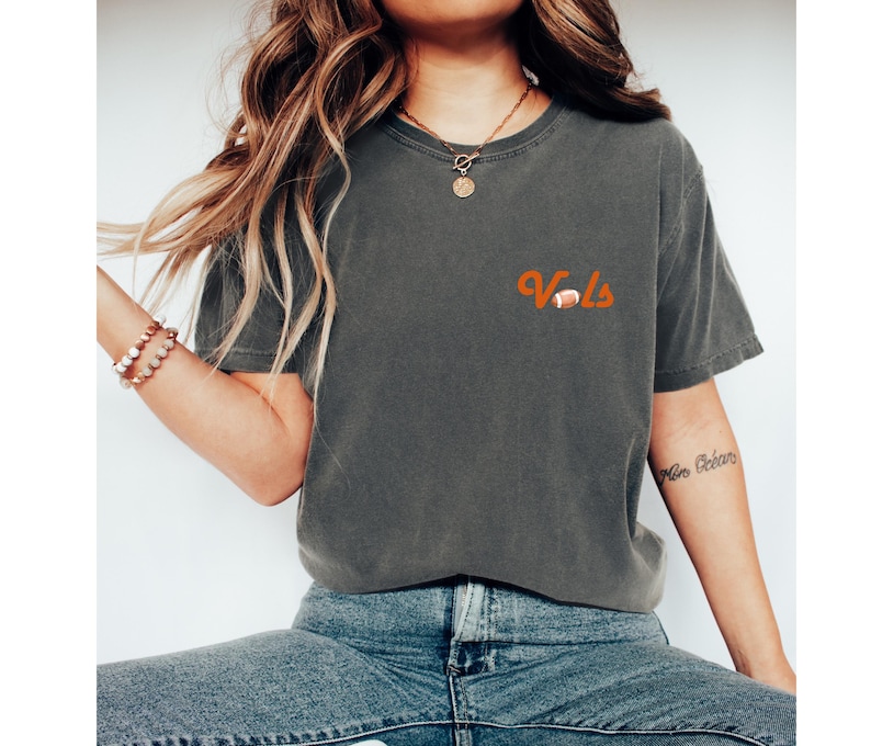 May include: A dark gray t-shirt with the word "Vols" in orange lettering and a small orange football graphic. The shirt is worn by a person with long brown hair.