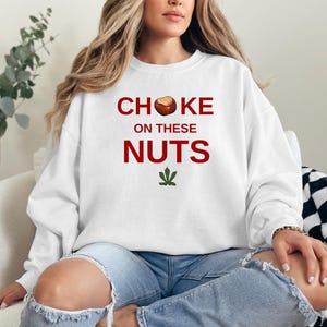 May include: White sweatshirt with the text "CHOKE ON THESE NUTS" in red letters and a brown nut graphic. A green marijuana leaf is below the text.