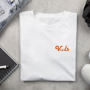 May include: White t-shirt with an orange graphic of the word "Vols" with a football.