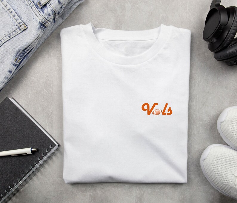 May include: White t-shirt with an orange graphic of the word "Vols" with a football.
