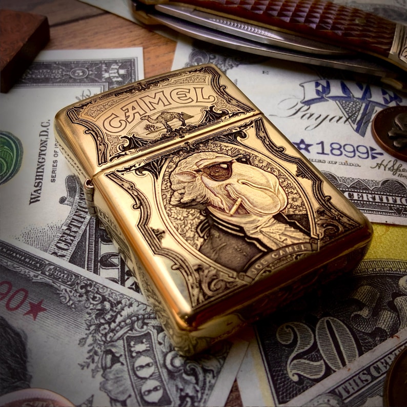 May include: A gold-colored metal lighter with intricate engraved details. The lighter features the word "CAMEL" and an image of a camel. The design includes a portrait of a man smoking a cigarette. The lighter is resting on a bed of US currency.