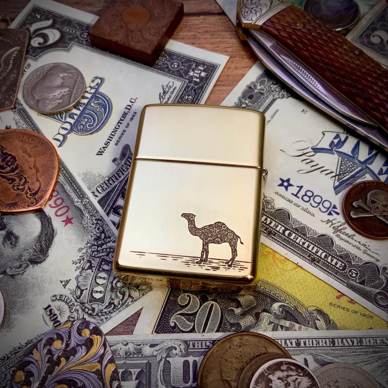 May include: A gold-colored metal lighter with a camel design etched on the front. The lighter is rectangular and has a shiny finish. The camel is standing on a line, possibly representing the desert. The background includes US currency, coins, and a pocket knife.