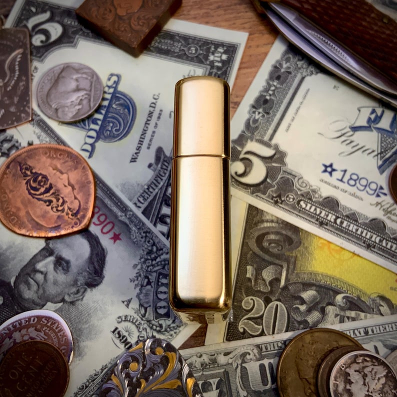 May include: A gold-colored, rectangular perfume atomizer stands upright on a surface covered with U.S. currency and antique coins. The atomizer has a cylindrical body and a cap. The background includes dollar bills and various coins.