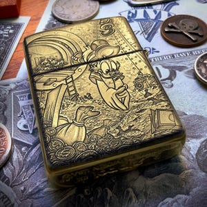 May include: A gold-colored metal lighter featuring a detailed engraved illustration of Donald Duck surrounded by money bags and coins. The design includes other cartoon characters and the number "3". The lighter is rectangular with rounded edges.